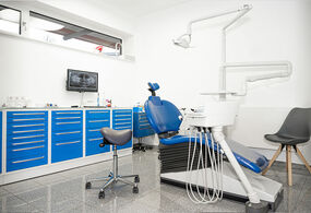 Ein Behandlungsraum in einer Zahnarztpraxis mit blauem Zahnarztstuhl, dunkelgrauem Hocker, Schränken mit blauer Front und Zahnarztgeräten. Auf der Arbeitsfläche auf den Schränken an der Wand steht ein Computermonitor und darüber gibt es ein Fenster. Der Raum ist hell und sauber.