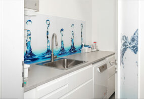 Eine Arbeitsfläche mit weißen Unterschränken und grauer Arbeitsplatte. Darin eingelassen befindet sich eine Edelstahlspüle. Die Rückwand zeigt ein künstlerisches Design aus Wassertropfen in Blautönen. Auf der Arbeitsfläche stehen ein paar kleine medizinische Utensilien.